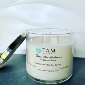 A scented candle labeled 'TAM' with a metal lid, set against a soft white backdrop.